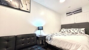 Ein Schlafzimmer mit einem Bett, einem schwarzen Ledersofa, einem Nachttisch mit Lampe und einer Wand-Klimaanlage.