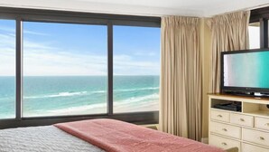 Ett hotellrum med ett stort fönster som vetter mot havet, en säng med ett rosa överkast och en tv-apparat som står på ett träbord.