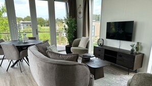 A modern living room with a sectional sofa, a coffee table, a TV, and a dining area with chairs and a table. - Just Nature Light 4 (Olburgen)
