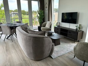 A modern living room with a sectional sofa, a coffee table, a TV, and a dining area with chairs and a table. - Just Nature Light 4 (Olburgen)
