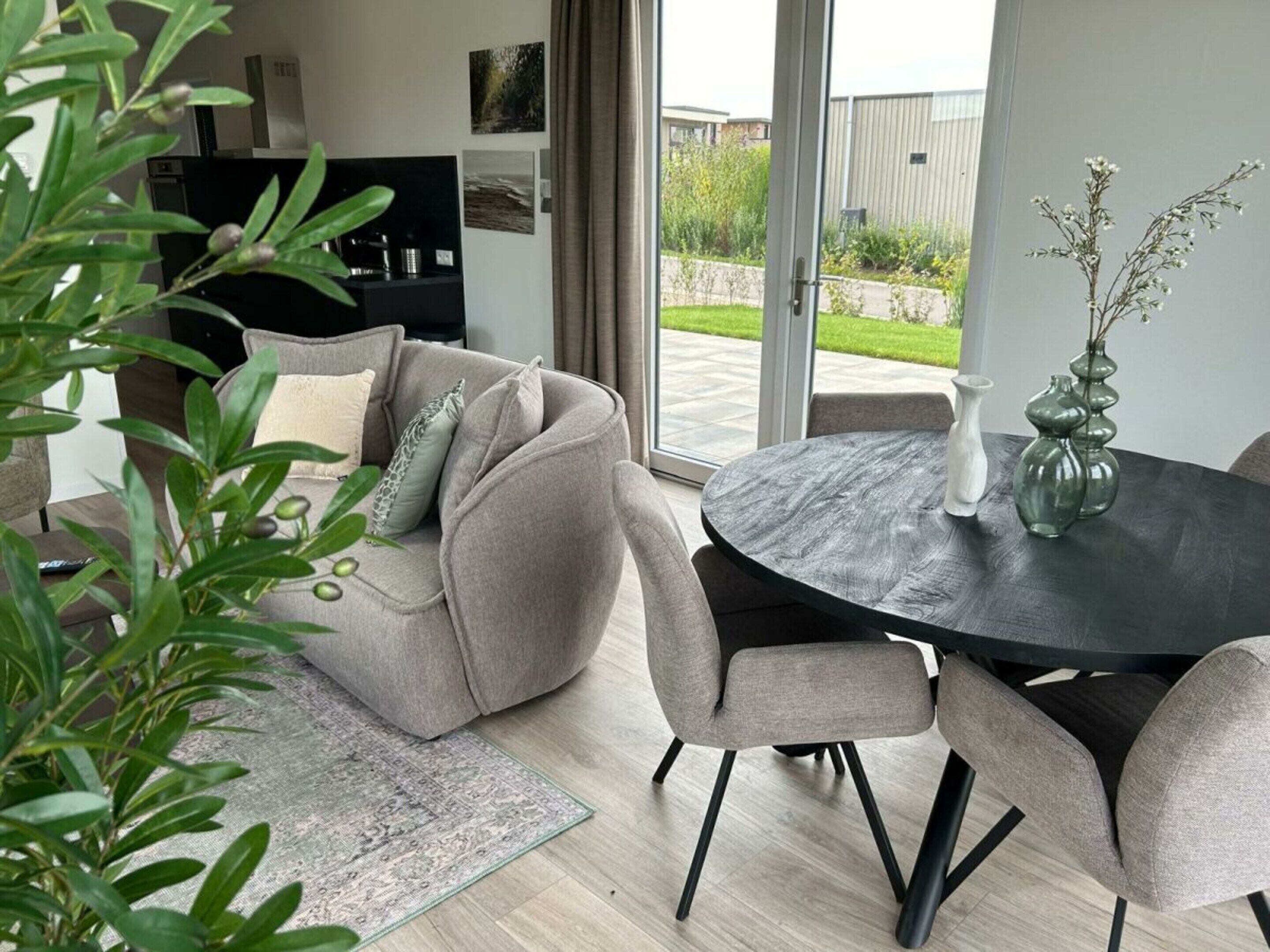 Ein modernes Wohnzimmer mit einem grauen Sofa, einem runden Tisch, Stühlen, einer Kaffeemaschine und Blick ins Freie.