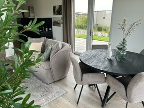 A modern living room with a grey sofa, a round table, and chairs, a coffee machine, and a view of the outdoors. - Just Nature Light 4 (Olburgen)