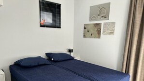 A room with two single beds, a window with blinds, a wall-mounted lamp, and a small table. - Just Nature Light 4 (Olburgen)