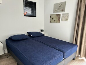 A room with two single beds, a window with blinds, a wall-mounted lamp, and a small table. - Just Nature Light 4 (Olburgen)