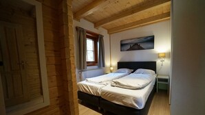Una habitación con techo de madera, una cama con sábanas blancas, una mesita de noche con una lámpara y una ventana con cortinas.