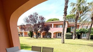 Eine Außenpatio mit Tisch und Stühlen, einem Fliesenboden und Blick auf ein Gebäude mit rosafarbenem Mauerwerk und Palmen.