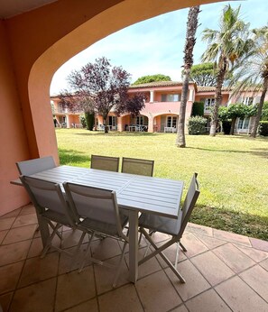 An outdoor patio with a table and chairs, a tiled floor, and a view of a building with a pink facade and palm trees. - Air-conditioned apartment with a heated pool, 10 minutes from Saint-Tropez (Grimaud)