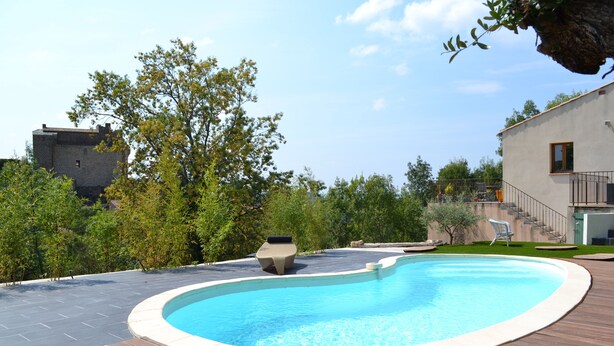 A swimming pool with a wooden deck, a stone planter, and a modern house in the background. - House with swimming pool, terraces and exceptional views. (Lunas-les-Châteaux)