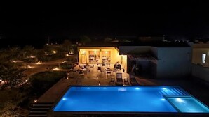 Una piscina con una zona de estar hundida, rodeada de un patio con mesas y sillas, y un edificio con un patio abierto al fondo.