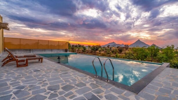 A swimming pool with stone flooring and metal railings, surrounded by greenery and a mountainous landscape during sunset.