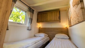 Un petit logement composé d’une seule pièce, comprenant deux lits, une fenêtre avec des rideaux, une tête de lit en bois et un meuble fixé au mur.