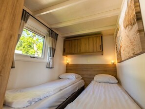 Eine kleine, einfache Unterkunft mit zwei Betten, einem Fenster mit Vorhängen, einem Holzkopfstück und einem an der Wand befestigten Schrank.