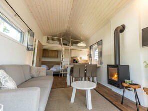 Ein gemütliches Interieur mit einer Couch, einem kleinen runden Tisch, einer Küchenzeile, einem Schlafzimmer auf Podest und einem Holzofen.