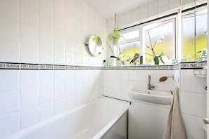 A bathroom with white tiles, a bathtub, a sink, a mirror, and potted plants. - The Guide's Charming Home (London)