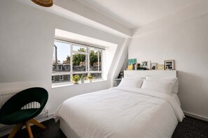Un dormitorio con una cama grande, una ventana con vistas a edificios, una silla y una mesita de noche con objetos.