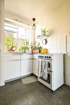 Cocina compacta con armarios blancos, estufa, fregadero y ventana con vistas a edificios.
