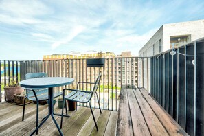 Una terrassa de fusta amb taula i cadires, una planta en test i una vista d'un edifici amb diverses finestres.