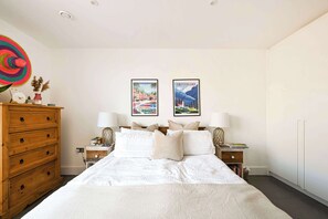 A bedroom with a large bed, wooden dresser, two bedside tables with lamps, and two framed pictures on the wall. - Modern Flat with Country Charm (London)