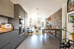 A modern kitchen with built-in appliances, a dining area with a brick wall, and a living room with a sofa and coffee table. - Modern Flat with Country Charm (London)