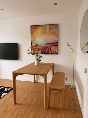 Ein Zimmer mit Holzboden, einem Tisch mit Bank, einem Bild an der Wand, einer Stehlampe und einem flachbildschirmfernseher, der an der Wand befestigt ist.