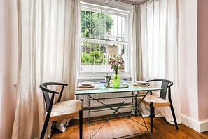 A dining area with a table and chairs, a window with curtains, and a radiator. - The Artist's Studio in Notting Hill (Greater London)