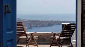 On aperçoit, à travers une porte bleue, deux chaises en bois et une table donnant sur une falaise et la mer.