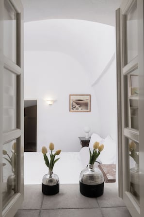 Un couloir menant à une chambre à coucher, où se trouvent un lit, un tableau encadré au mur et deux vases avec des fleurs.