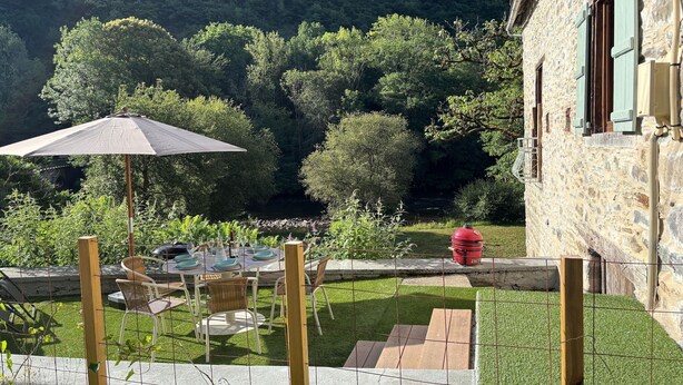 A stone building with a fenced patio area, outdoor seating, and a view of a forested hillside. - Château de Comborn Riverside Gite with Private River Access in the Vézère Valley (Orgnac-sur-Vézère)