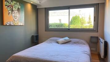 A bedroom with a large window, a bed, a painting on the wall, and a radiator.