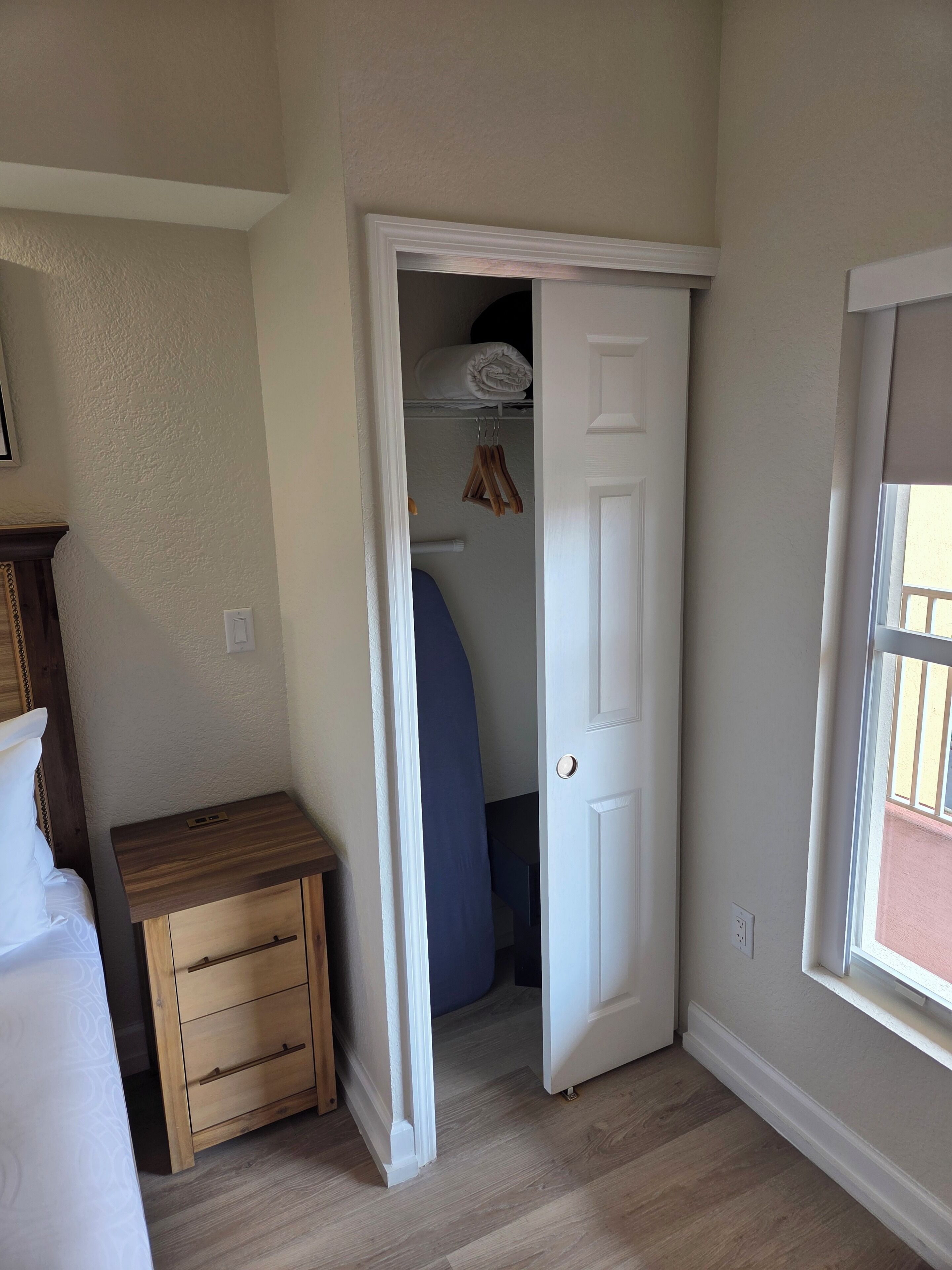 Une chambre à coucher avec une table de chevet en bois, un lit, une armoire où sont suspendus des vêtements et une fenêtre avec des stores.