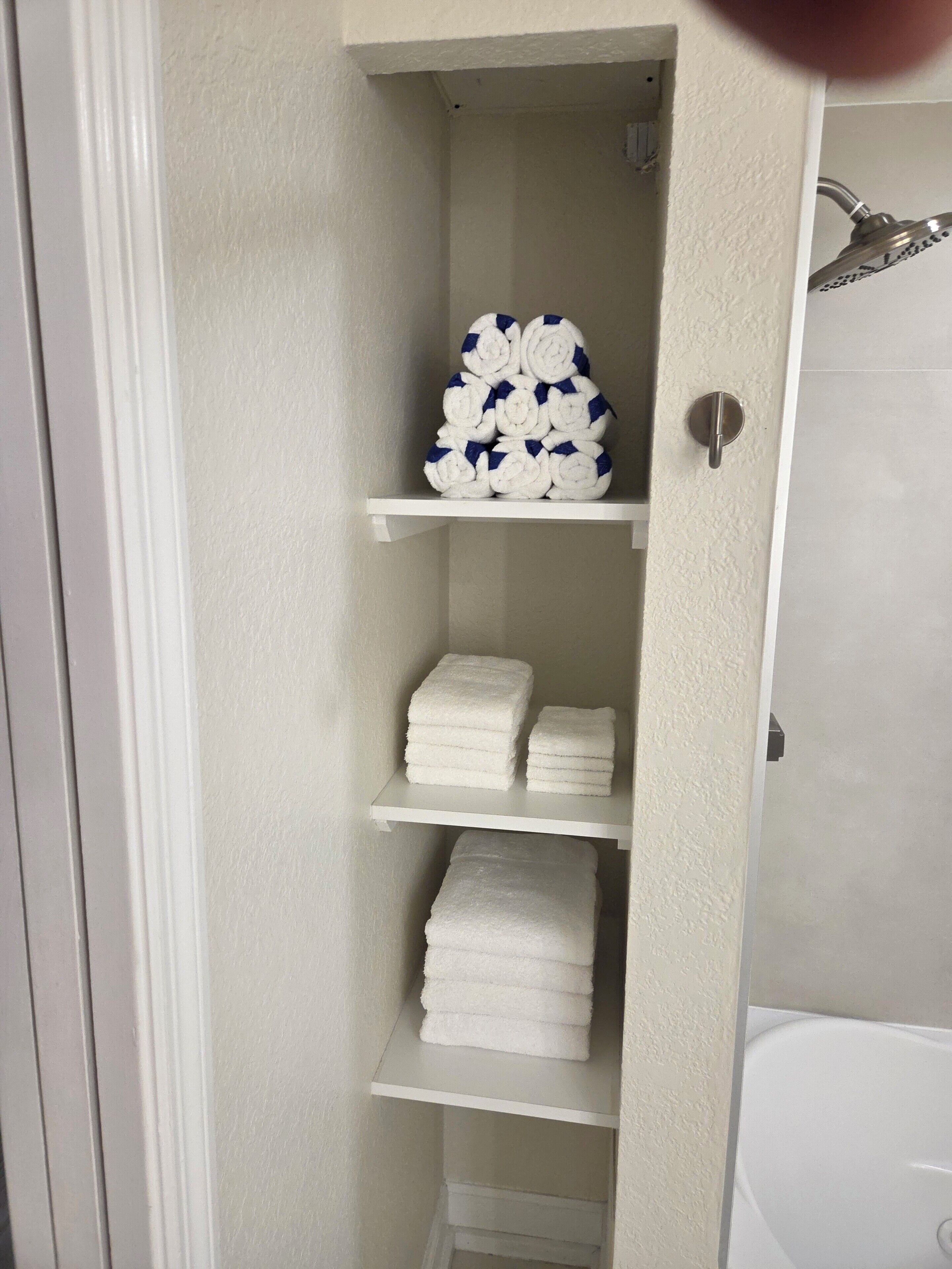 Une salle de bain avec une baignoire blanche, une douche dotée d’un pommeau de douche argenté et des étagères où sont rangées des serviettes blanches.