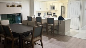 Un comedor con mesa y sillas de madera, una cocina con muebles blancos, un microondas y un refrigerador.