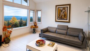 A living room with a sofa, a painting, a vase with flowers, and a view of trees through large windows. - Contemporary Bellevue Home with Panoramic Lake & Mountain Views (Bellevue)