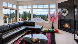A modern living room with a sectional sofa, a glass coffee table, a fireplace, and large windows offering a view of trees and a cloudy sky. - Contemporary Bellevue Home with Panoramic Lake & Mountain Views (Bellevue)