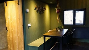 Una habitación con puerta de madera, un banco, una mesa con sillas, una planta colgante y una ventana con persianas.