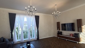 A spacious living room with a large window, two chandeliers, a flat-screen TV, and a wooden entertainment unit. - Spacious 4-bedroom home with air conditioning in beautiful Baku near Miami Beach (Baku)