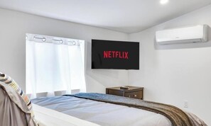 A bedroom with a bed, a TV mounted on the wall, a small table, and an air conditioning unit.