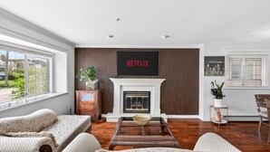 A living room with a fireplace, a large TV mounted above it, and a view of the outdoors through a window. - Modern 6BR |Near Downtown| Fast WiFi| Free Parking (Vancouver)