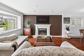 A living room with a fireplace, a large TV mounted above it, and a view of the outdoors through a window. - Modern 6BR |Near Downtown| Fast WiFi| Free Parking (Vancouver)