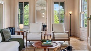 A room with two white armchairs, a small round table with books, a lamp, and a view of greenery through large windows. - Pool + Deck Dining + BBQ | Kayaks + EV Charger • Catskills View | River View (Germantown)