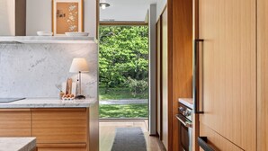 A modern kitchen with marble countertops, wooden cabinets, a built-in stove, and a view of a green garden. - Pool + Deck Dining + BBQ | Kayaks + EV Charger • Catskills View | River View (Germantown)