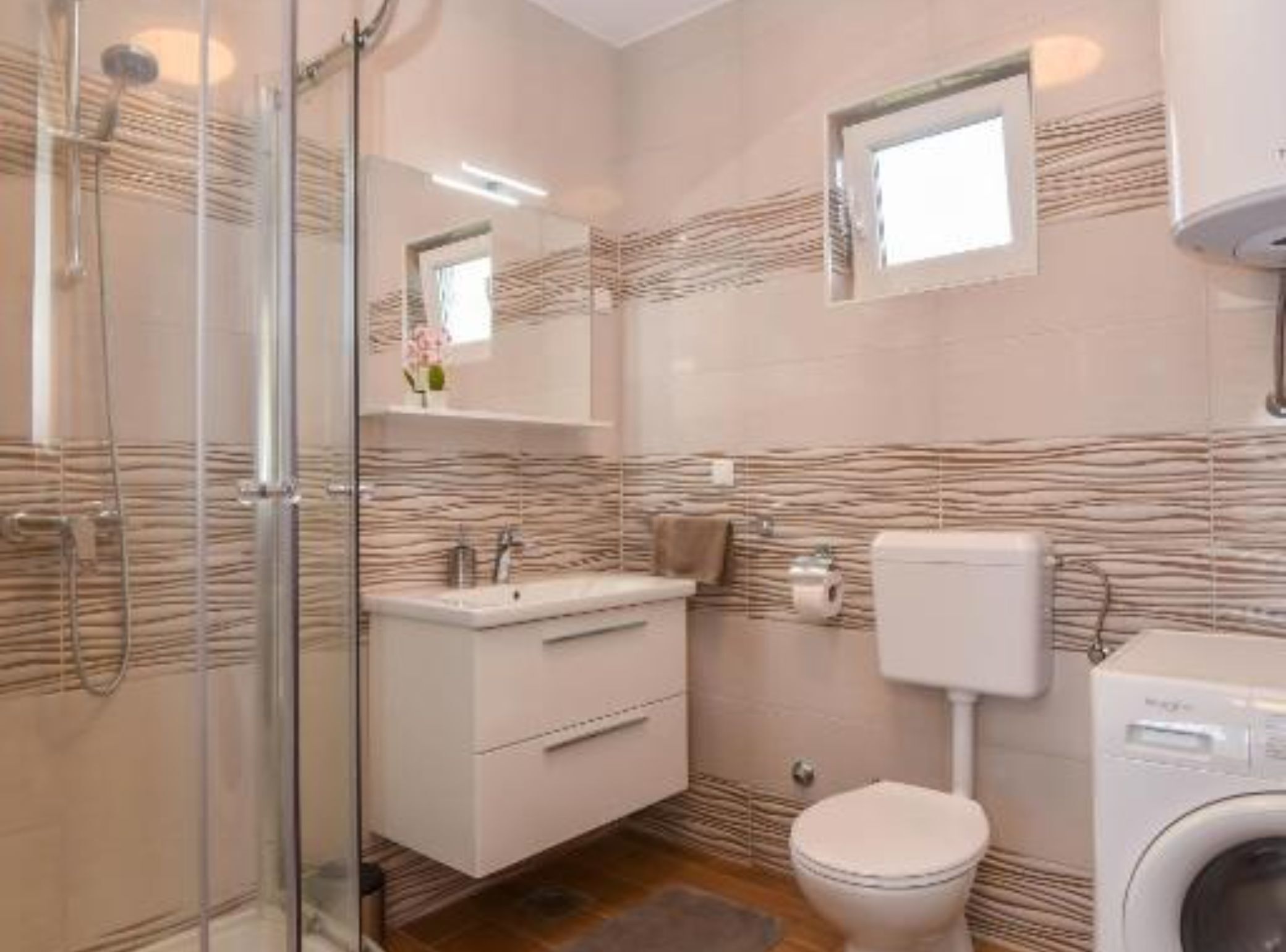 A bathroom with a glass shower enclosure, a sink with a mirror, a toilet, and a washing machine.
