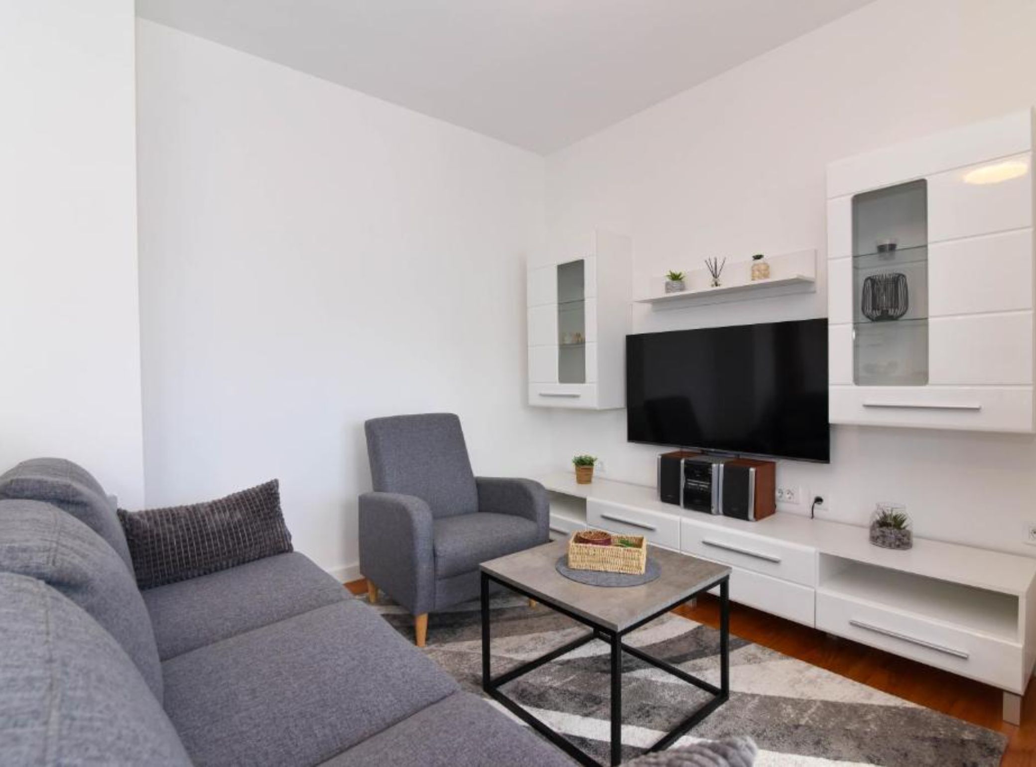 A modern living room with a grey sofa, a matching armchair, a coffee table, a flat-screen TV, and built-in cabinets.