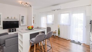 Una sala moderna con un televisor de pantalla plana, una pequeña isla de cocina con un frutero, dos taburetes grises, un sofá gris y un ventanal con cortinas.