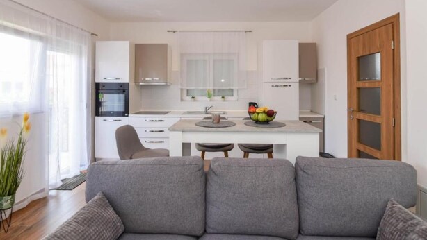 Una cocina moderna con armarios blancos, una isla central con frutero y una zona de comedor con sofá gris.