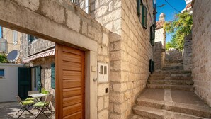 Un bâtiment en pierre avec une porte en bois, un petit espace extérieur aménagé avec une table et des chaises, et un escalier en pierre menant à un niveau supérieur.