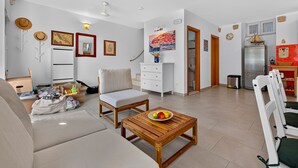 Un salon comprenant un canapé, une table basse en bois avec des fruits, une chaise blanche, un meuble blanc et un coin cuisine avec un réfrigérateur et un élément de décoration rouge sur le mur.