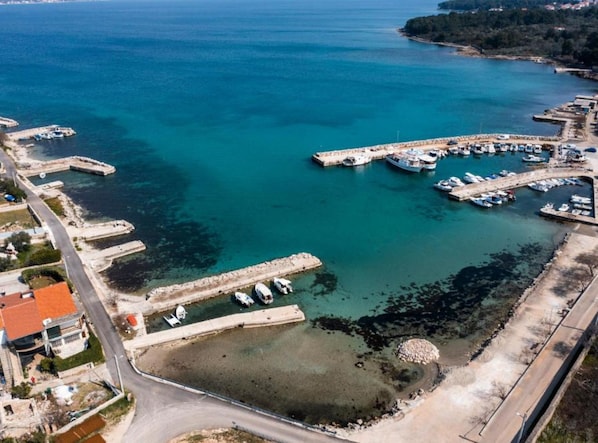 Luftaufnahme eines Küstenhafens mit vertäuten Booten, einer kleinen Insel und einer Straße, die zum Hafen führt.