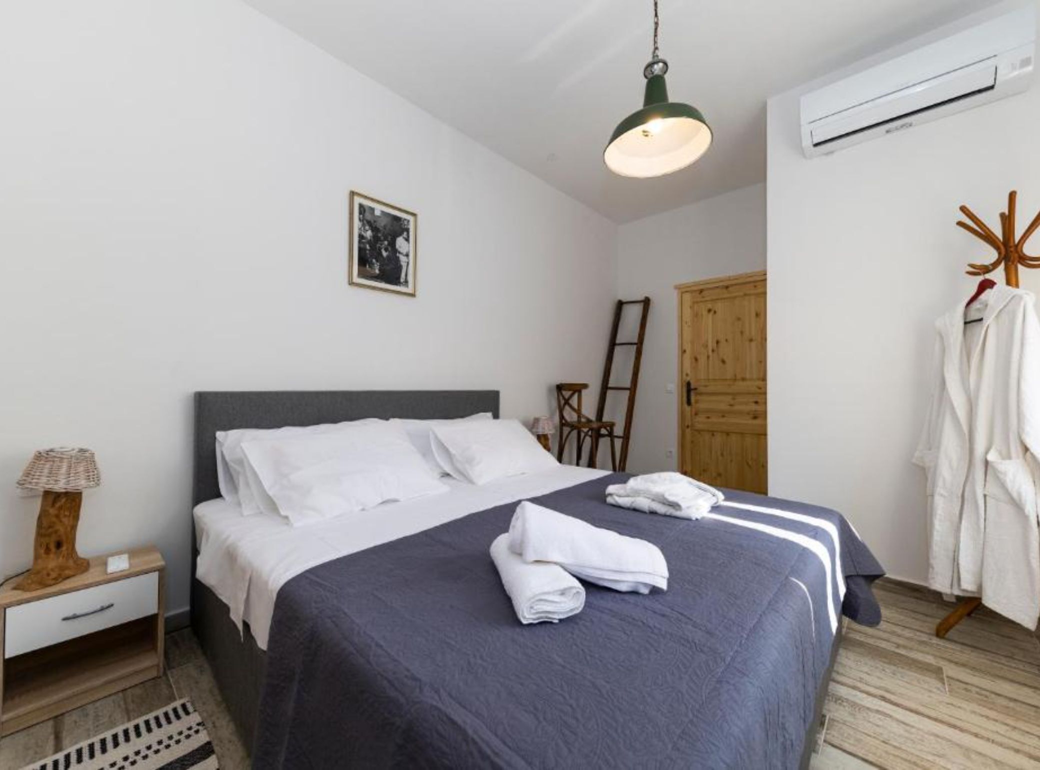 Un lit bien fait, avec une literie blanche et bleu foncé, une échelle en bois, une armoire en bois et un peignoir suspendu.