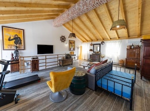 Ein geräumiger Wohnbereich mit Holzboden, einem Laufband, einem gelben Sessel, einem blauen Sofa und einem an der Wand befestigten Fernseher.